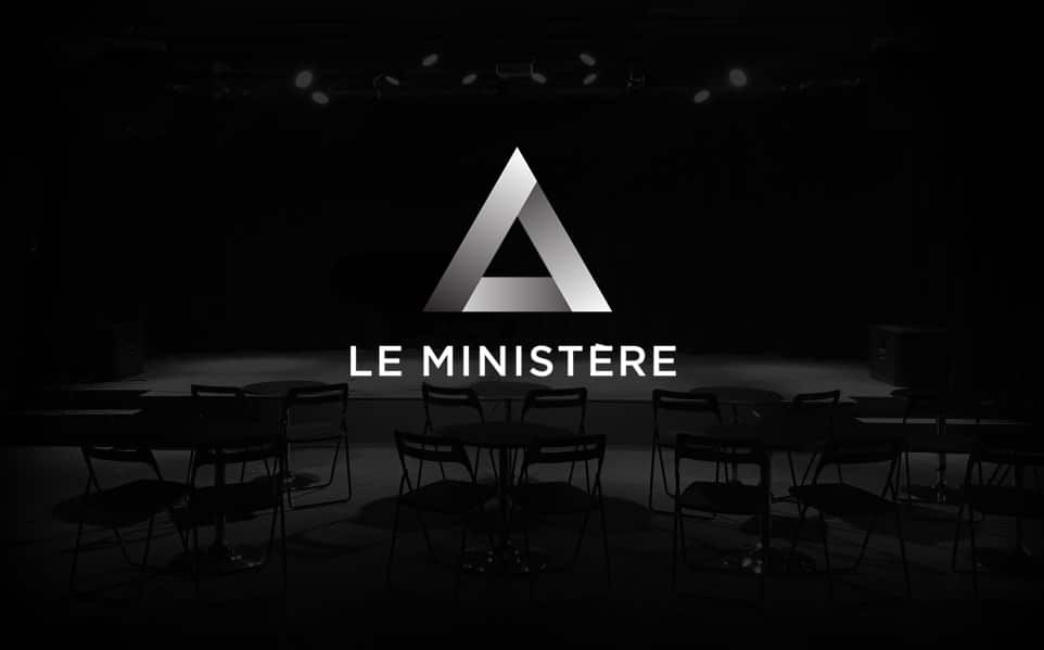 Une salle sombre avec des chaises et des tables vides, éclairée par des projecteurs. Au centre, un logo triangulaire métallique est affiché au-dessus du texte 